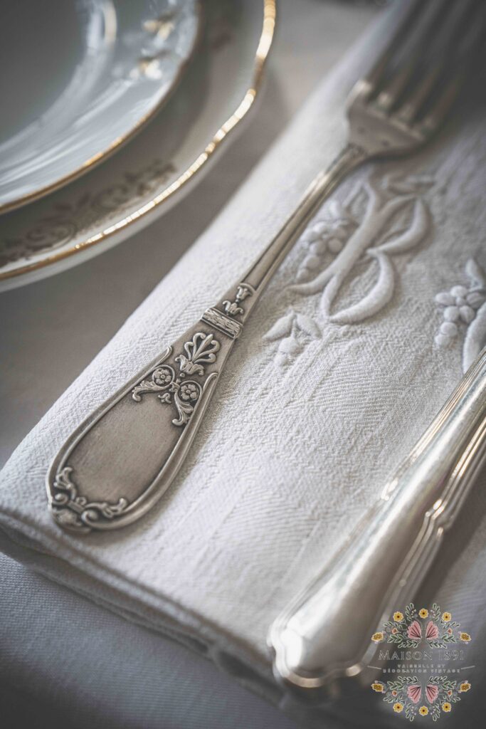 Vue de dessus d'une serviette ancienne brodée en lin blanche, repliée. Posés dessus des couverts en métal argentés ornementés. Location de linge ancien Lyon - Location vaisselle et décoration vintage - Maison 1891